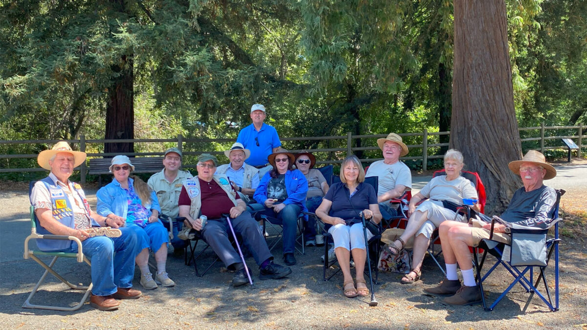 Members of Santa Clara Valley Carvers and their significant other are sitting and chatting under large redwood trees