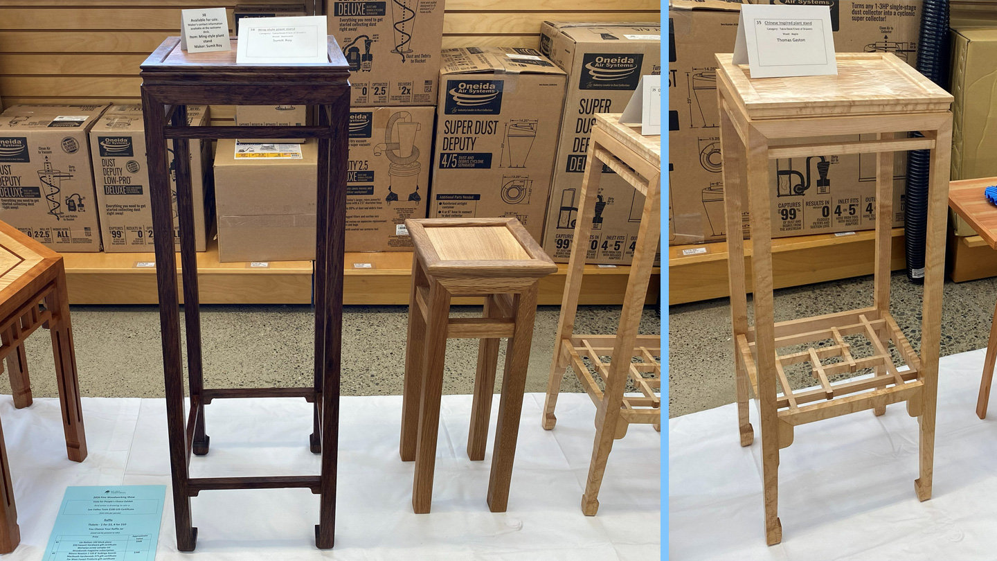 Several handmade wooden plant stands and small tables are displayed on a white cloth at a woodworking show at Woodcraft San Carlos. On the left, a tall, dark-stained stand features a square top and traditional joinery. Next to it is a shorter, medium-toned wooden stand. To the right, a tall, light-colored stand showcases a square top and a distinctive geometric lattice stretcher near the bottom. Small information cards rest on the tops of the furniture pieces.