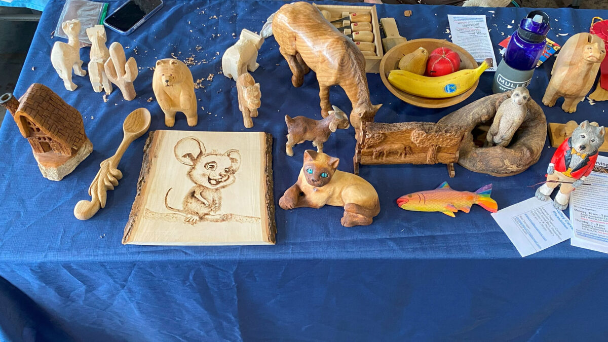 A table with woodcarving and woodburning projects, including a carved and painted rainbow fish.