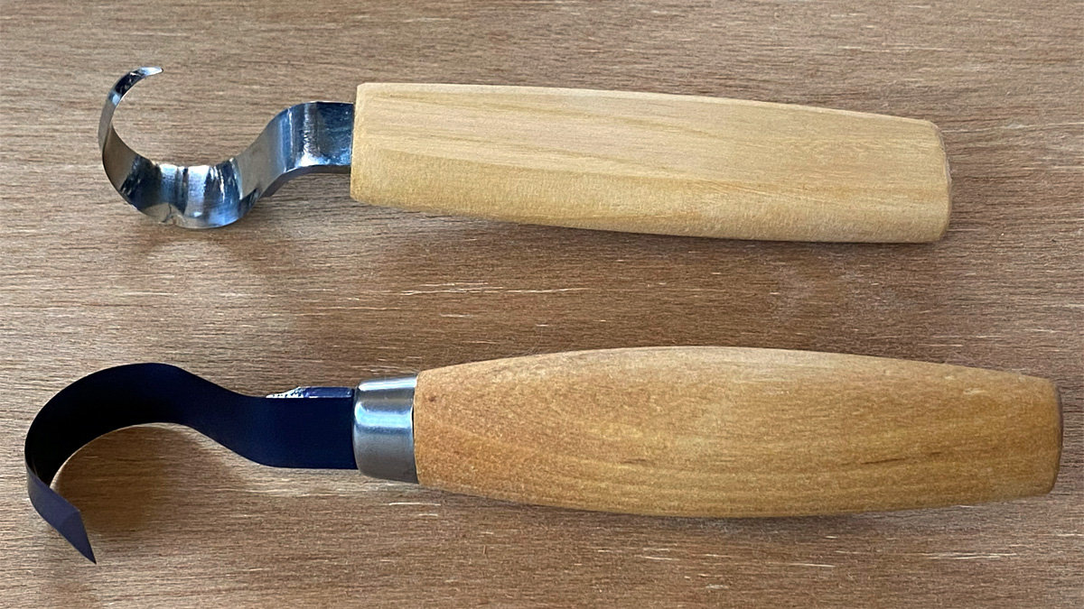 Two woodcarving hook knives with wooden handles lying horizontally side-by-side on a light wood surface. The top tool features a shiny, silver-colored, curved blade and a light, blocky handle without a ferrule. The bottom tool has a dark blue or blued steel curved blade with a pointed tip, separated from its medium-brown rounded wooden handle by a metal collar.