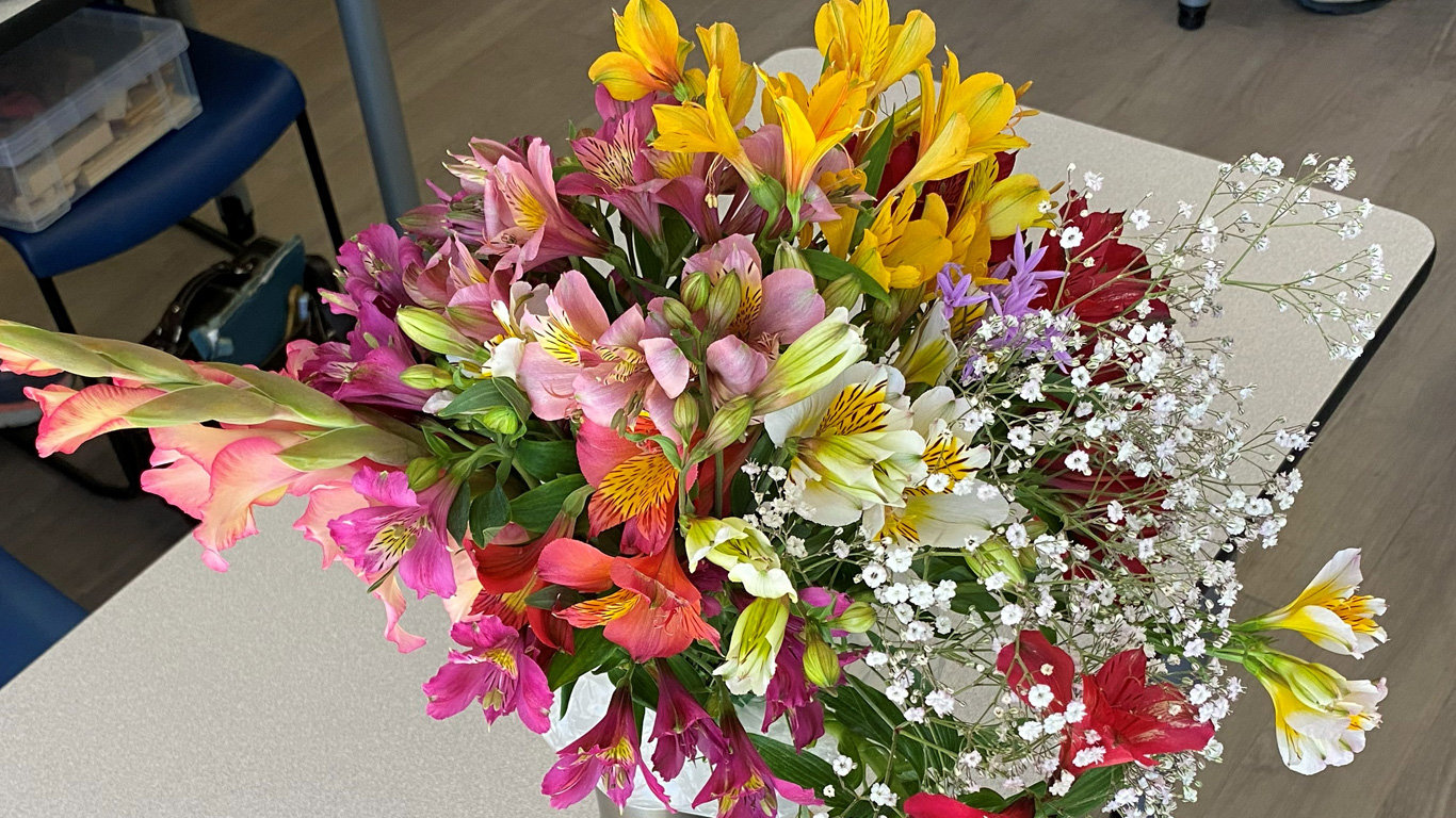 A large, vibrant bouquet of spring flowers sits on a light-colored table. The arrangement features numerous Alstroemeria (Peruvian lilies) in shades of bright yellow, magenta, pink, and white with yellow-striped throats. A tall stalk of peach and pink gladiolus extends from the left side of the bouquet, while delicate sprays of tiny white baby's breath fan out on the right.