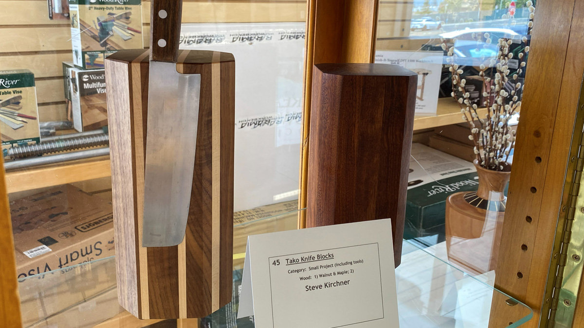 Two faceted wooden knife blocks are displayed on a glass shelf. The taller block on the left is made of alternating vertical strips of dark walnut and light maple wood, with a large cleaver attached to the front. To its right sits a shorter, solid dark walnut block of a similar geometric shape. A white card in the foreground identifies the pieces as "Tako Knife Blocks" by Steve Kirchner.