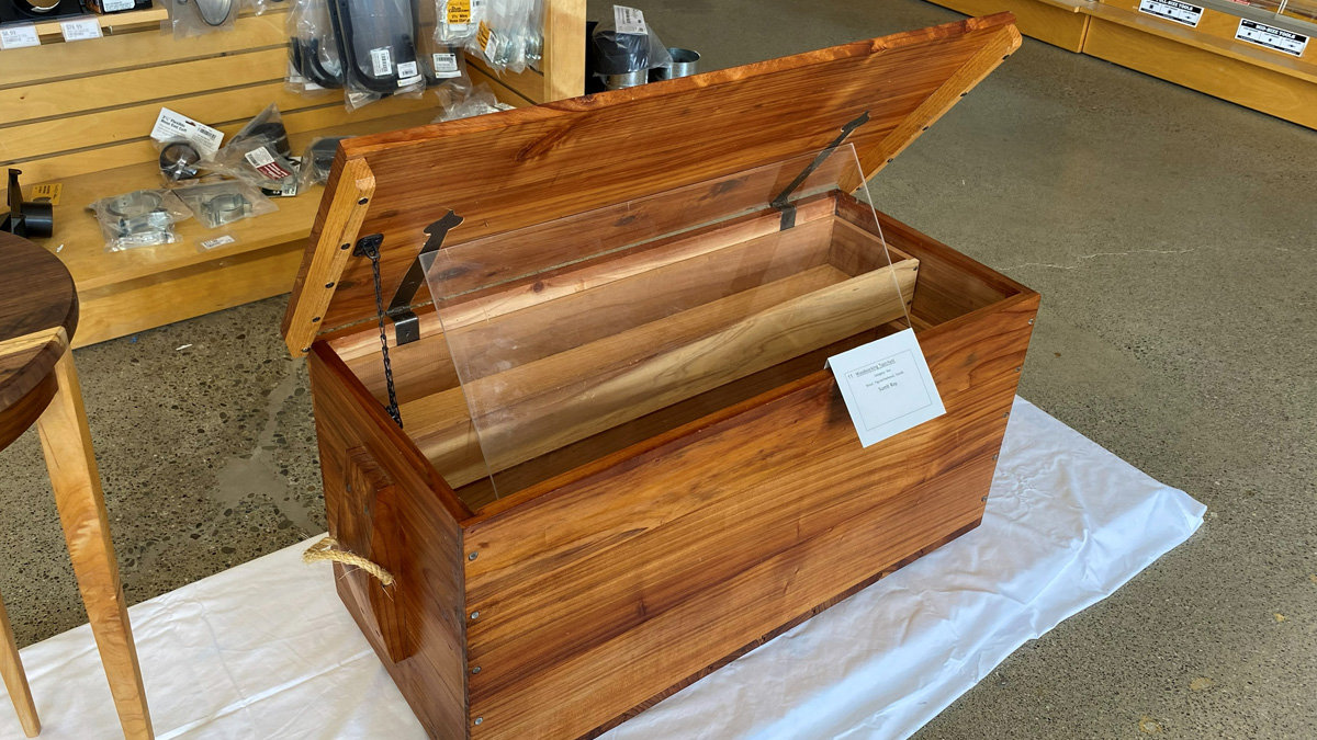 A wooden tool chest with a rich, reddish-brown finish is displayed with its lid propped open. Inside, a sliding tray made of lighter-colored wood is visible. The chest features black metal hinges, a chain mechanism to hold the lid up, and a rope handle attached to the side. A small white information card is attached to the front right corner.