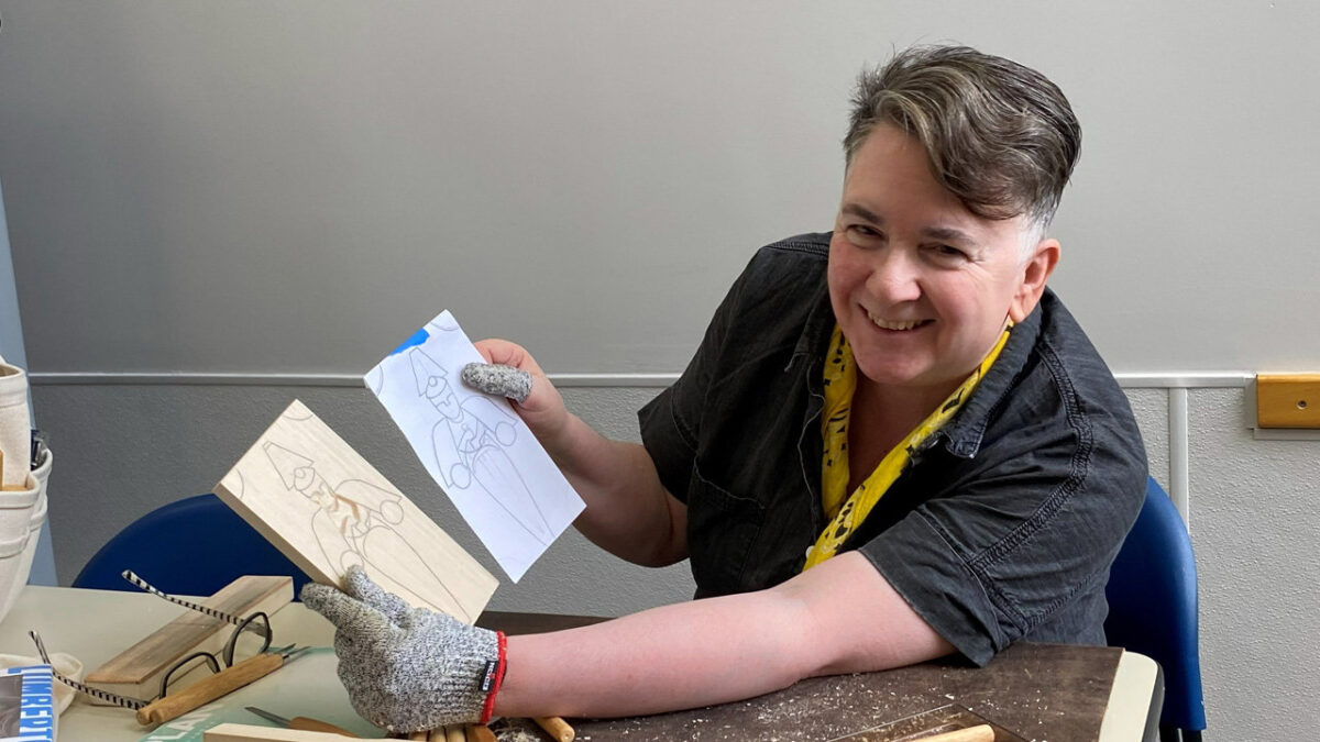 Terry Davis smiles while holding up a block of carved basswood next to its original pencil sketch design.