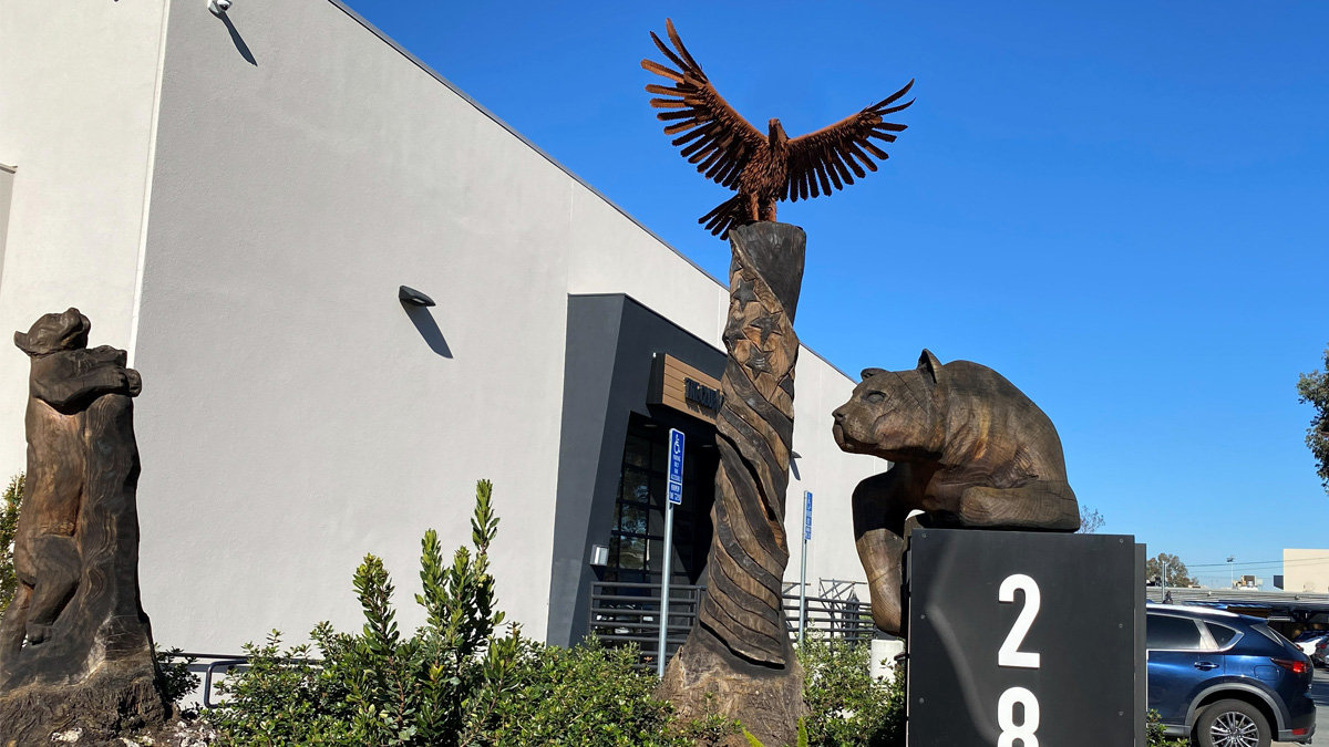Large wooden sculptures displayed outdoors featuring a standing bear leaning against a post, a tall twisted pole carved with stars and topped by an eagle, and a large reclining mountain lion resting on a pedestal.