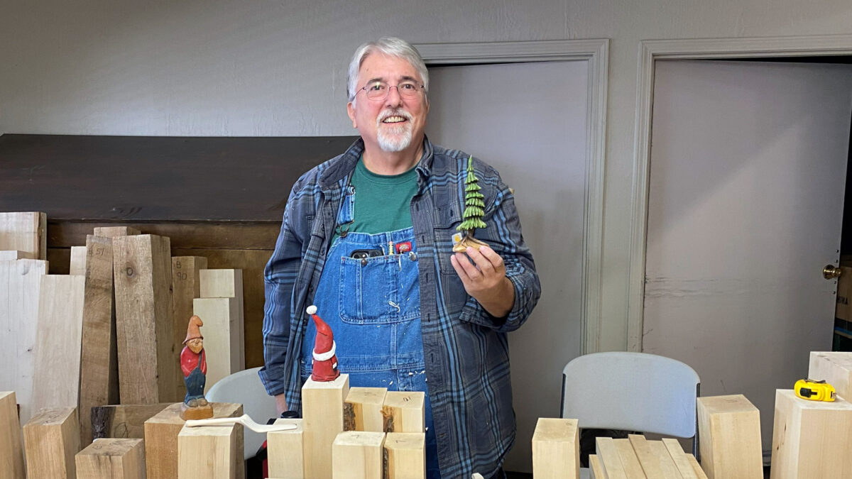 Tom Checketts stands smiling behind a table stacked with raw lumber blocks at the Oakhurst Woodcarvers Rendezvous, wearing denim overalls and holding up a small wooden carving of a green tree in his left hand. On the table around him are various wood blocks and other small carvings, including a gnome figure standing on a block to the left and a small Santa in a red coat and hat resting on a stack near the center.