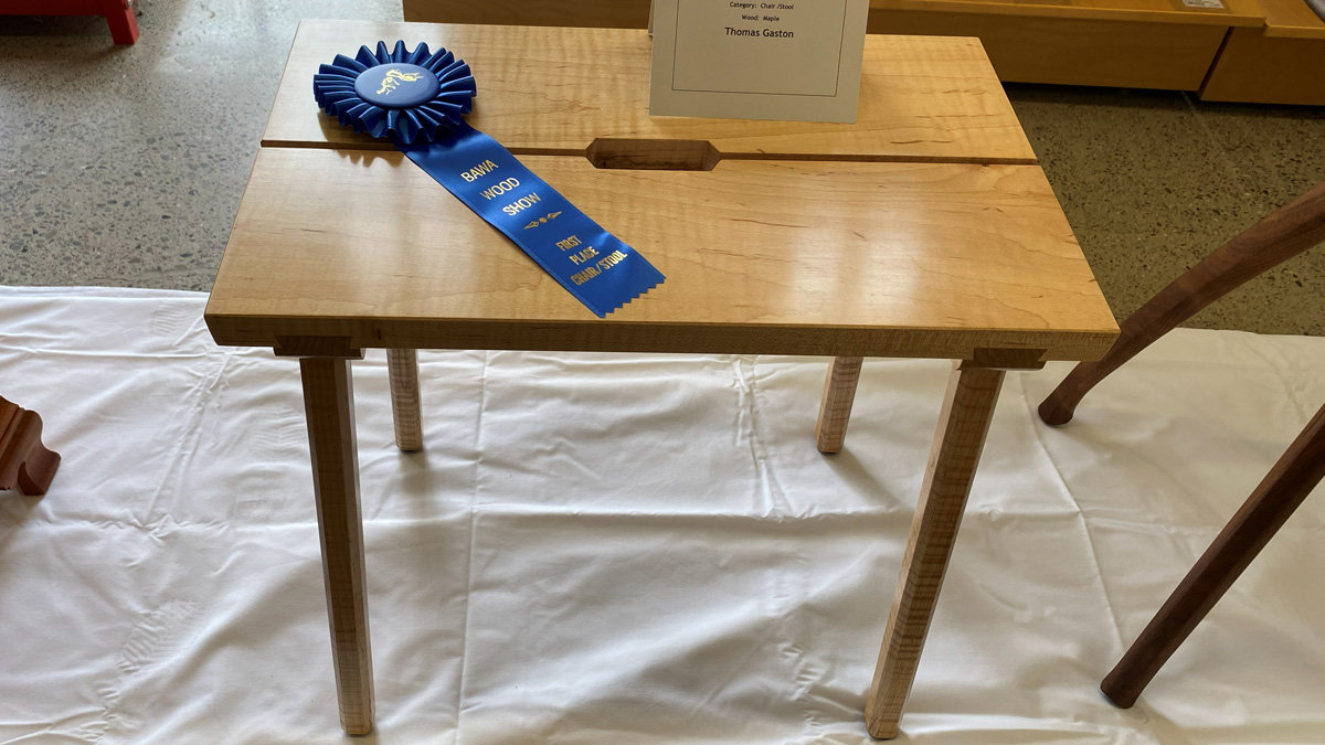 A light-colored maple stool with a rectangular top constructed from two joined slabs featuring a central joinery slot. The stool stands on four straight, square legs attached to an apron beneath the seat. A blue award ribbon labeled "BAWA Wood Show - First Place Chair/Stool" rests on the left side of the top, next to a white card identifying the maker as Thomas Gaston.