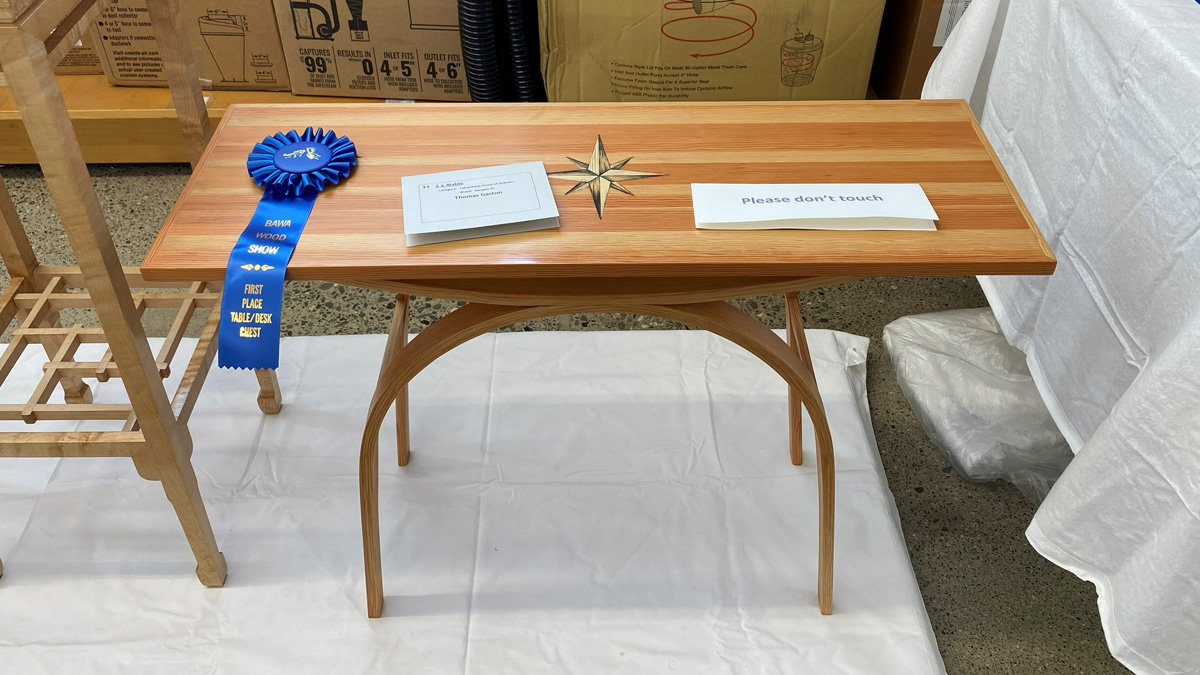 A light-colored wooden table featuring a geometric, eight-pointed star inlay in the center of the tabletop. The table has distinctive, curved legs that arch outward. A blue ribbon attached to the left side reads "RAVA Wood Show First Place Table/Desk Chest." A small white card in the center identifies the project as "2 x 4 Table," implying it was built from a single 2x4 Douglas Fir piece of wood. The table was constructed by the maker Thomas Gaston.