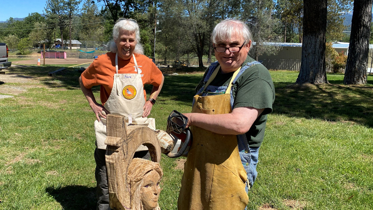 At the 2026 Oakhurst Woodcarvers Rendezvous, woodcarver Vic Hood poses outdoors with fellow carver Ann Bruner-Welch. Ann stands on the left wearing an orange shirt and a beige apron with her name written on it, smiling with hands on her hips. Beside her, Vic wears glasses, a green shirt over denim overalls, and a heavy tan leather apron while holding a chainsaw. Between them is a large block of wood featuring a partially completed sculpture that includes an archway and the detailed face of a woman.