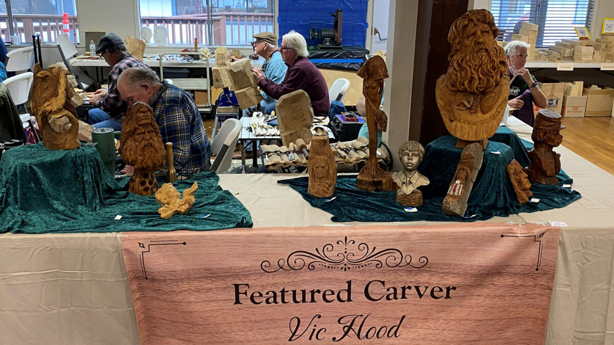A display table features the work of woodcarver Vic Hood at the Oakhurst Woodcarvers Rendezvous in April 2026. A banner reading "Featured Carver Vic Hood" hangs from the front, while teal cloths on the table showcase various wooden sculptures, including detailed busts of a mountain man, a young boy, and other figures. In the background, attendees can be seen working on projects at tables in a community hall with large windows looking out onto a deck.