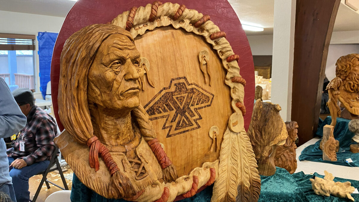 This large wooden relief carving by artist Vic Hood features a detailed bust of an elderly Native American man with braided hair set against a circular background resembling a ceremonial drum or shield. In the center of the light-colored wood, a geometric Thunderbird symbol is rendered in dark pyrography, surrounded by a rope-textured rim and hanging carved feathers on the right side. The artwork rests on a table covered with teal cloth at the Oakhurst Woodcarvers Rendezvous in April 2026.