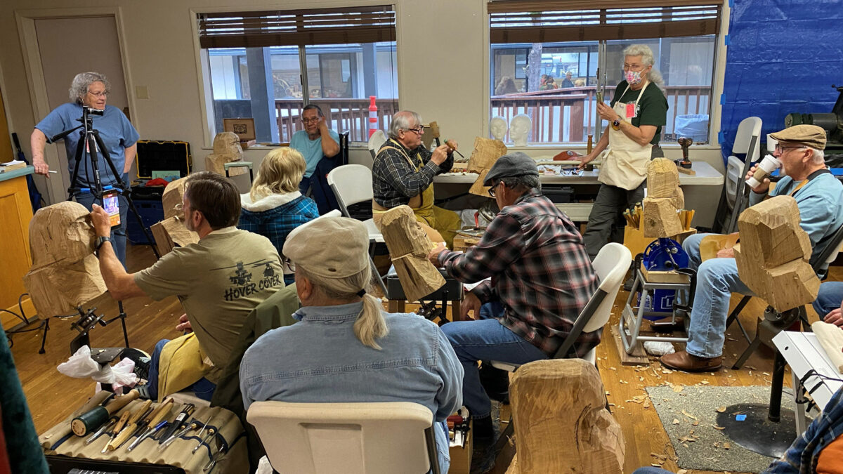 During a bust-carving workshop at the 2026 Oakhurst Woodcarvers Rendezvous, several students sit working on wooden head sculptures clamped to stands. In the foreground, men in casual attire carve rough blocks of wood, while instructor Vic Hood stands in the background using a chisel with a mallet. Wood shavings are scattered across the floor, and a case of carving chisels is open in the bottom left corner.