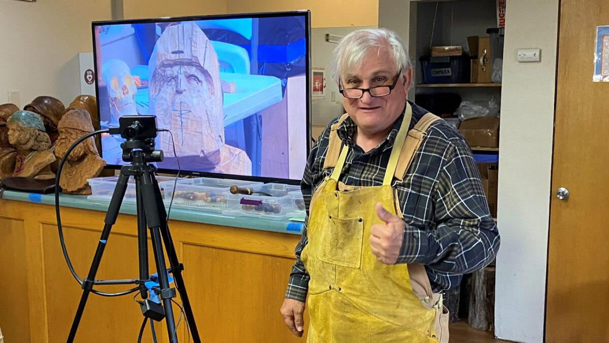 Vic Hood, wearing a yellow leather apron over a plaid shirt, stands in a workshop giving a thumbs-up gesture next to a black camera mounted on a tripod. Behind him, a large television screen displays a close-up image of a wooden bust carving, while several other completed wooden profile sculptures sit on the counter to the left amidst shelves of supplies.