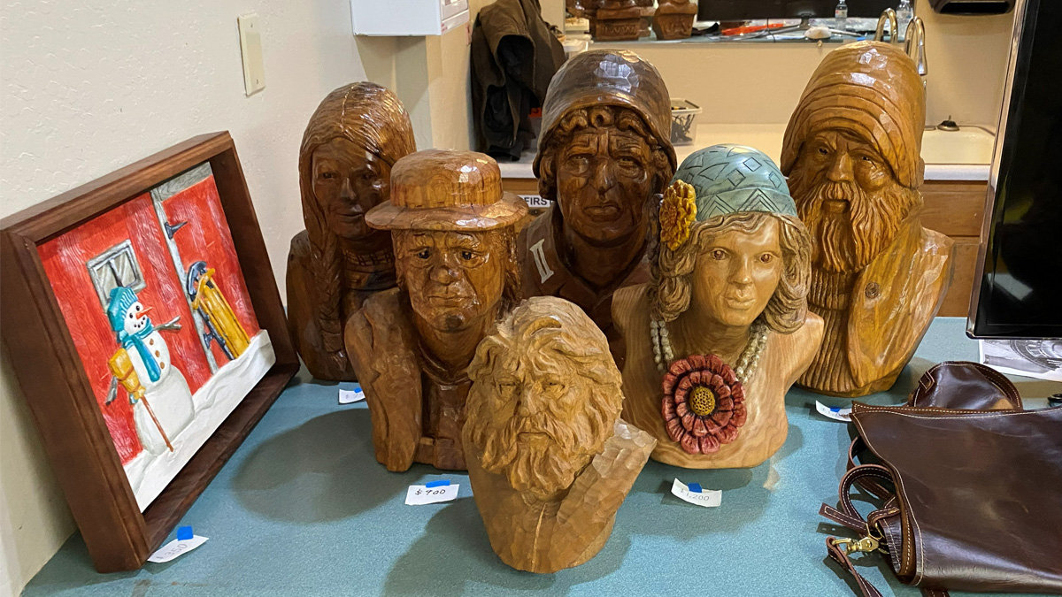 A collection of wooden sculptures displayed on a teal counter at Vic Hood’s booth for the 2026 Oakhurst Woodcarvers Rendezvous features a framed snowman relief carving alongside six carved busts, including a stern man in a flat cap, an elderly mountain man with a turban, and a young woman styled as a 1920s flapper wearing a blue hat and large red flower brooch.