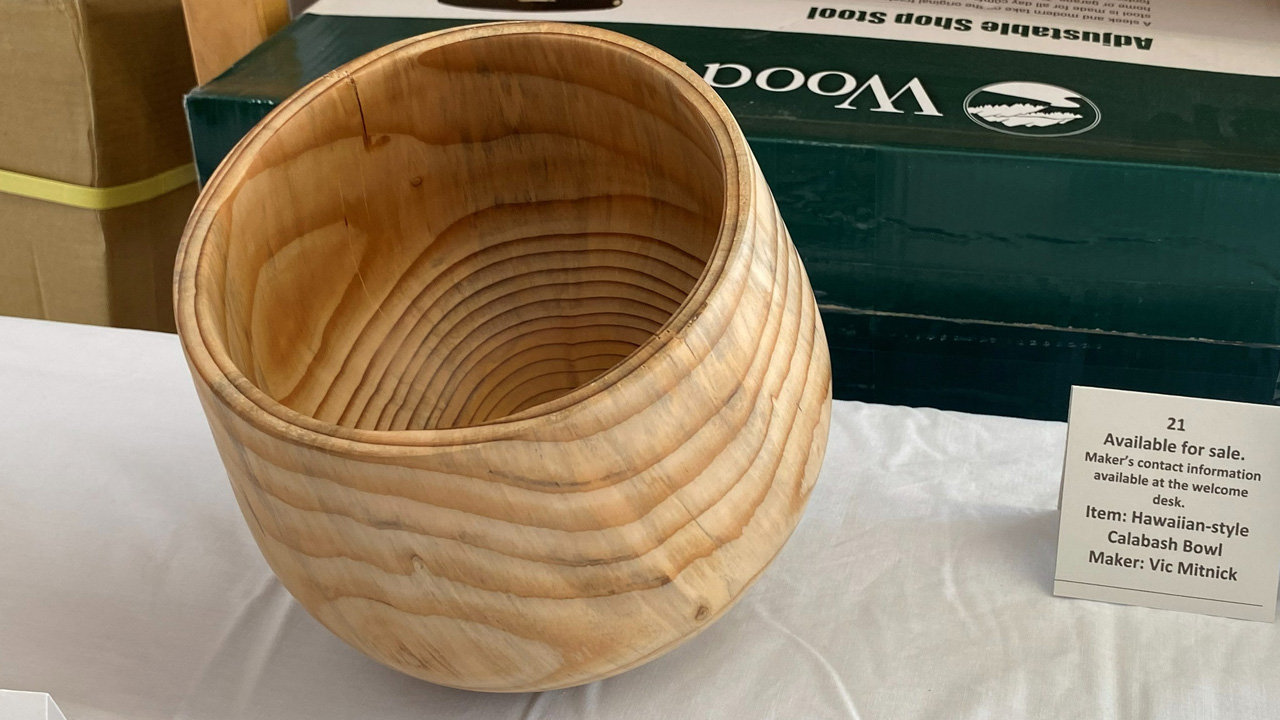 A light-colored, hand-turned wooden bowl from Norfolk Island Pine by maker Vic Mitnick, identified by a nearby placard as a "Hawaiian-style Calabash Bowl." The bowl features a rounded, bulbous body that tapers slightly toward a wide, flared rim. The exterior surface and the hollow interior clearly display the natural, concentric growth rings of the wood.