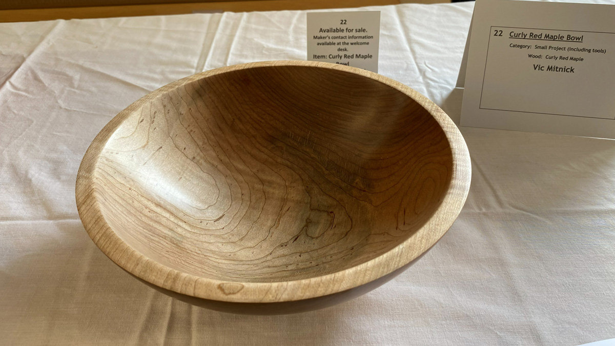A turned wooden bowl made of curly red maple by Vic Mitnick. The bowl features a light tan, honey-colored wood with distinct, wavy grain patterns (figure) running horizontally across the interior and exterior. It has a smooth, rounded rim and a shallow, open shape with a satin finish.