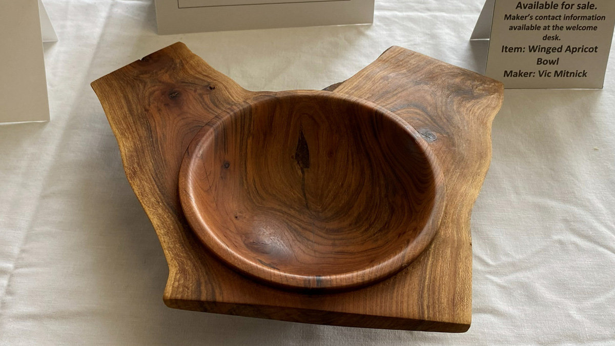 A wooden "Winged Apricot Bowl" crafted by Vic Mitnick. The piece features a polished, round bowl hollowed out of the center of a larger, irregular wooden slab. Two wing-like extensions flank the bowl, preserving the wood's natural live edge. The grain is prominent, showing swirling reddish-brown patterns throughout the piece.