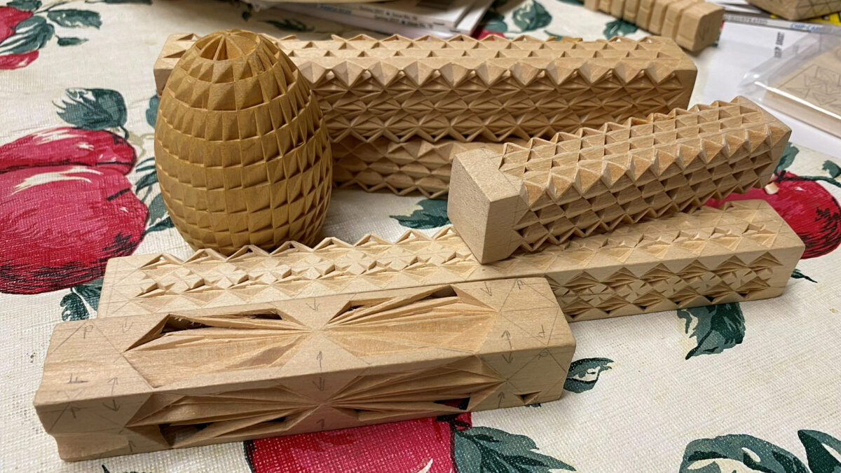 A close-up view of several chip carving projects resting on a tablecloth with a red apple pattern. The collection, carved by Vickie Hopson, features a large, textured wooden egg and multiple rectangular blocks intricately carved with geometric patterns of triangles and pyramids. In the foreground, a block shows pencil markings with arrows and letters outlining a starburst design, illustrating the layout stage of the carving process, while the pieces behind it display completed, textured surfaces. The items were on display at the Oakhurst Woodcarvers Rendezvous in April 2026.
