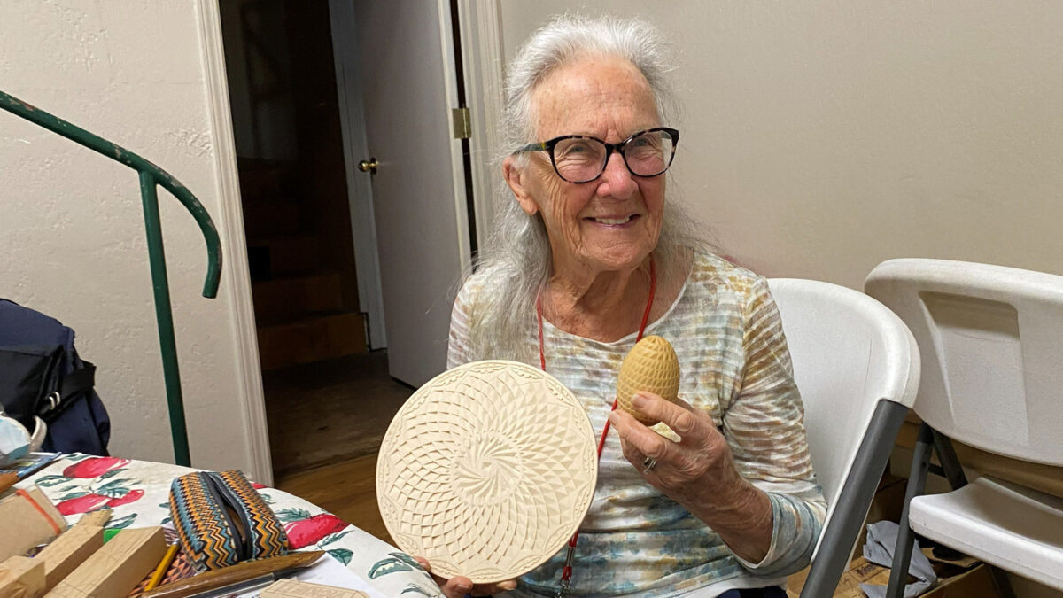 A smiling older woman with long white hair and glasses, Vickie Hopson, sits at a table covered with a floral cloth, holding up two chip-carving projects. In her right hand, she holds a flat, a wooden platter with an intricate geometric spiral pattern carved into the light-colored wood (basswood). In her left hand, she holds a small, wooden egg with a textured cross-hatch pattern. Woodcarving tools and basswood blocks are visible on the table beside her.