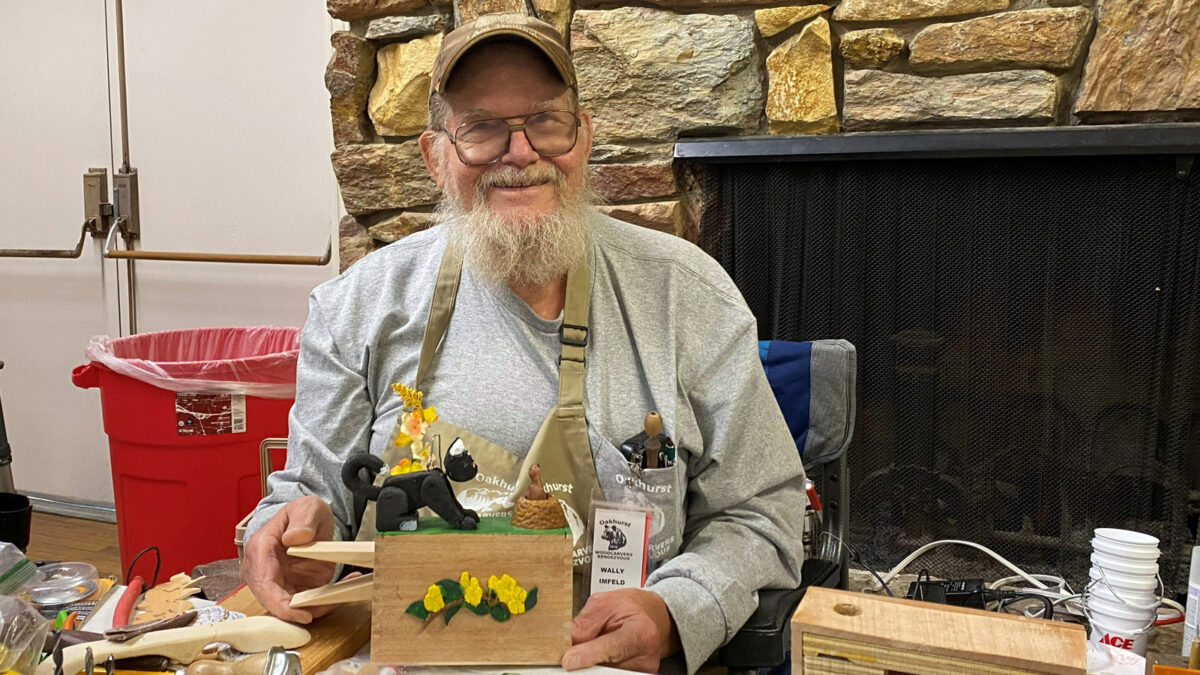An older man with a white beard and glasses, identified by his name tag as Wally Imfeld, smiles while demonstrating a wooden automata project at a workbench. He wears a beige apron filled with tools over a grey sweatshirt and holds onto wooden dowels attached to the side of a small box. The light-colored wooden box is decorated on the front with carved yellow flowers and green leaves. On top of the box, resting on a green surface, are small figures including a black cat and a gopher emerging from a mound, illustrating his mechanical carving style at the Oakhurst Woodcarvers Rendezvous.