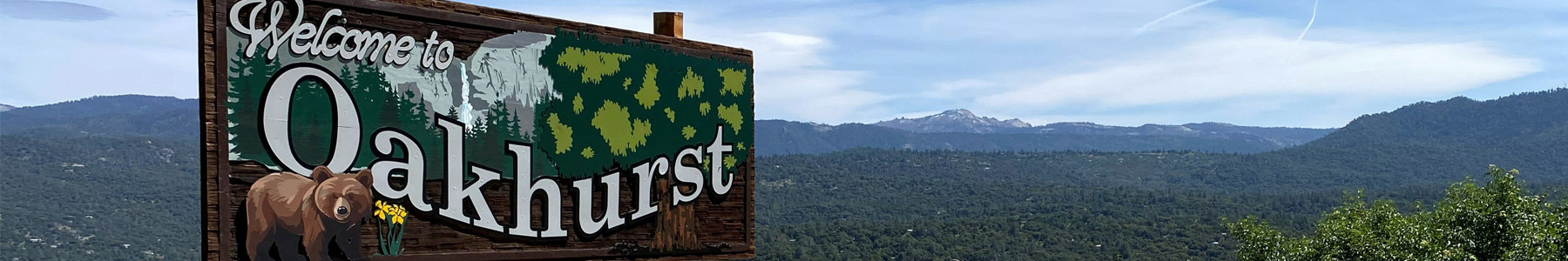 A large, handcrafted wooden sign reading "Welcome to Oakhurst" stands in the foreground on the left, featuring detailed carvings and paintings of a brown bear, evergreen trees, and rocky cliffs. Behind the sign is a scenic panorama of rolling, forest-covered mountains stretching toward a distant peak beneath a blue sky dotted with wispy clouds. The sign is located near Yosemite National Park in Oakhurst, California.