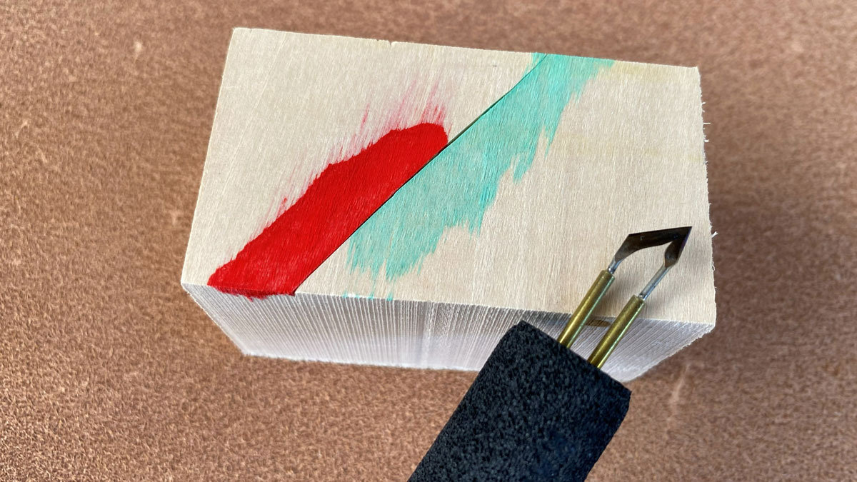Wood block with a clean cut separating two painted sections, bright red on one side, light green on the other, showing a sharp color line. Small paint bleed is visible where the paint touches the unpainted grain. A woodburning tip rests beside the cut, illustrating how to separate fibers before painting.