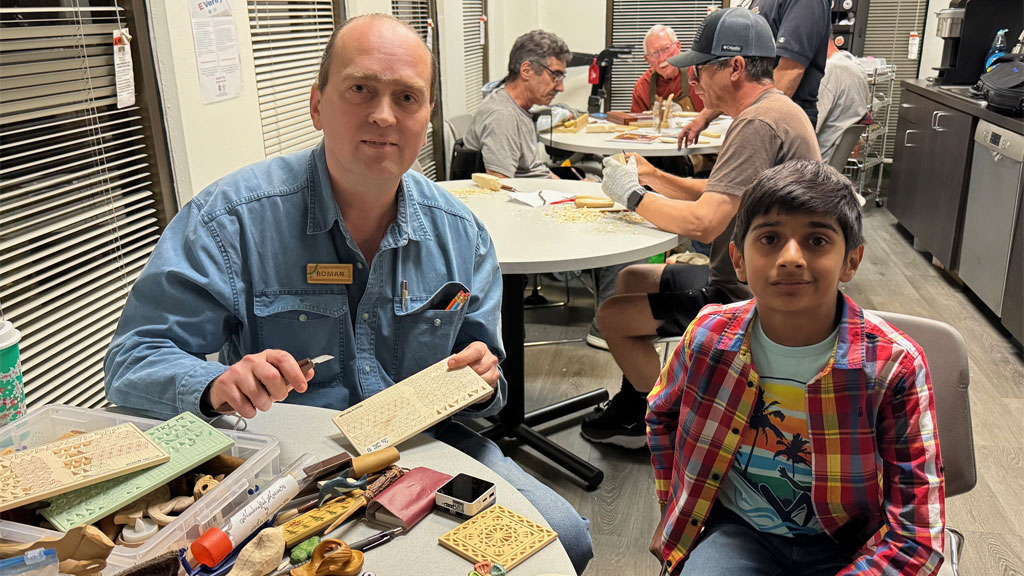 Roman shows woodcarving projects and tools to a guest. The club members carve in the background.