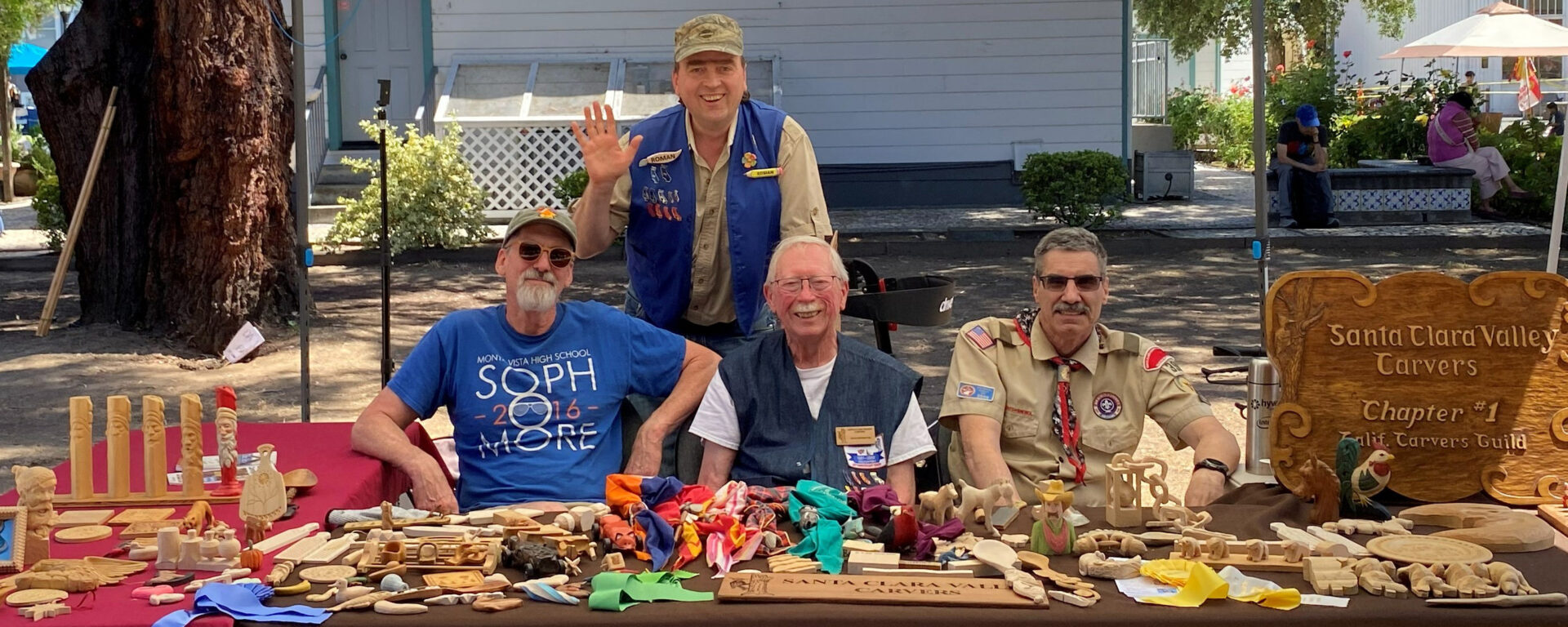 Four men pose at an outdoor booth for the Santa Clara Valley Carvers during a Scout-O-Rama event. Three men sit at a table covered with various woodcarvings, including totem poles, small animals, and wooden toys, while a fourth man stands behind them, waving. A large wooden sign on the right reads "Santa Clara Valley Carvers Chapter #1 California Carvers Guild." The man on the far right (Mike Campi) wears a tan scout leader uniform, and the man standing (Roman Chernikov) wears a blue vest with woodcarving enamel pins. Two other volunteers are David Clarke and David Osterlund.