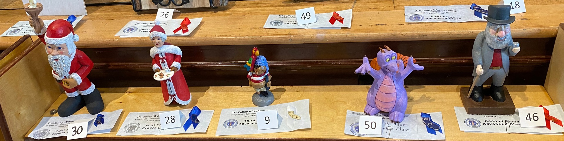 Award-winning wooden carvings of figures like Santa Claus and a purple dragon displayed with prize ribbons and entry cards at the Tri-Valley Carvers Show.