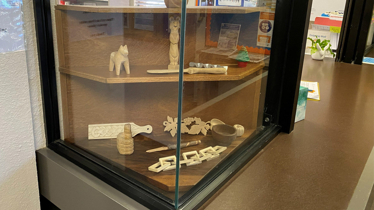 Glass display case featuring various woodcarvings, including animal figures, a standing bear, a Celtic knot coaster, grape-leaf spoon, a small bowl, and a wooden chain.