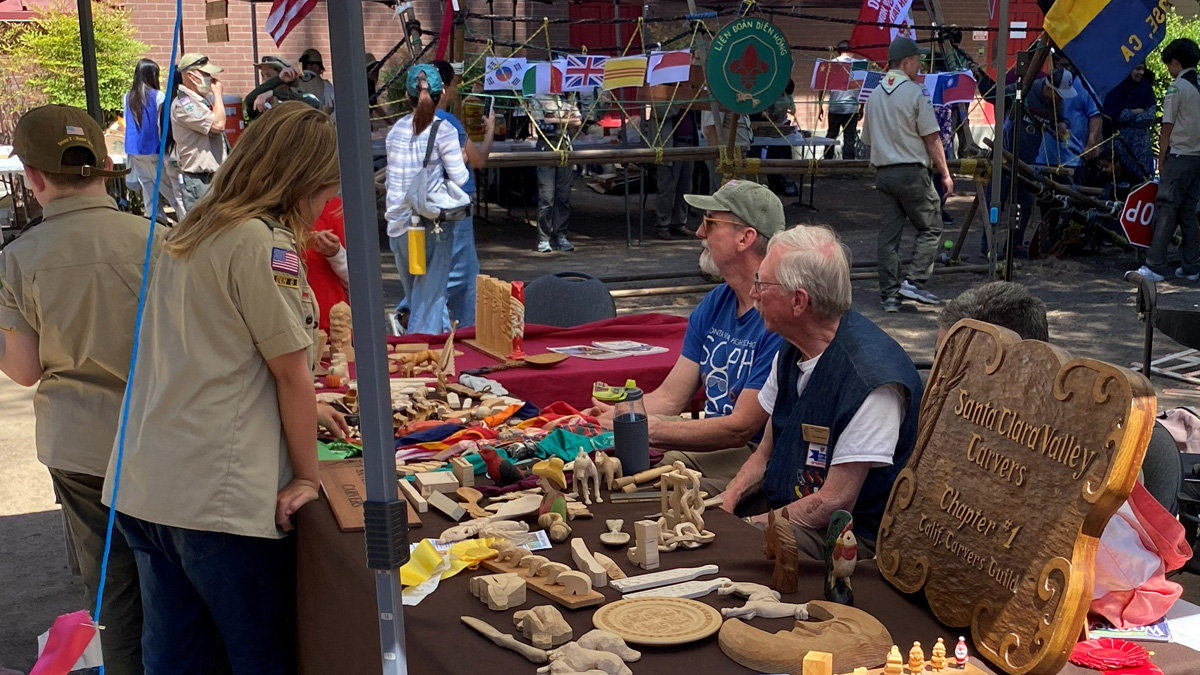 At an outdoor event called Scout-O-Rama held at History Park in San Jose, members of the Santa Clara Valley Carvers manage a booth filled with wooden crafts. In the foreground, a table covered in a brown cloth displays an extensive collection of woodcarvings, including chess pieces, small animal figurines, and turning tools. A large, ornate wooden sign on the right identifies the group as "Santa Clara Valley Carvers, Chapter #1 of California Carvers Guild". Three men (David, Dave, and Mike) sit behind the table, demonstrating woodcarving. They are interacting with a young woman with long blonde hair wearing a tan scout uniform, who is browsing the items. To the left, another scout in uniform stands with their back to the camera. In the background, beneath a canopy, a string of international flags hangs above other attendees and scouts, including a green circular sign with Vietnamese text, indicating a multicultural gathering.