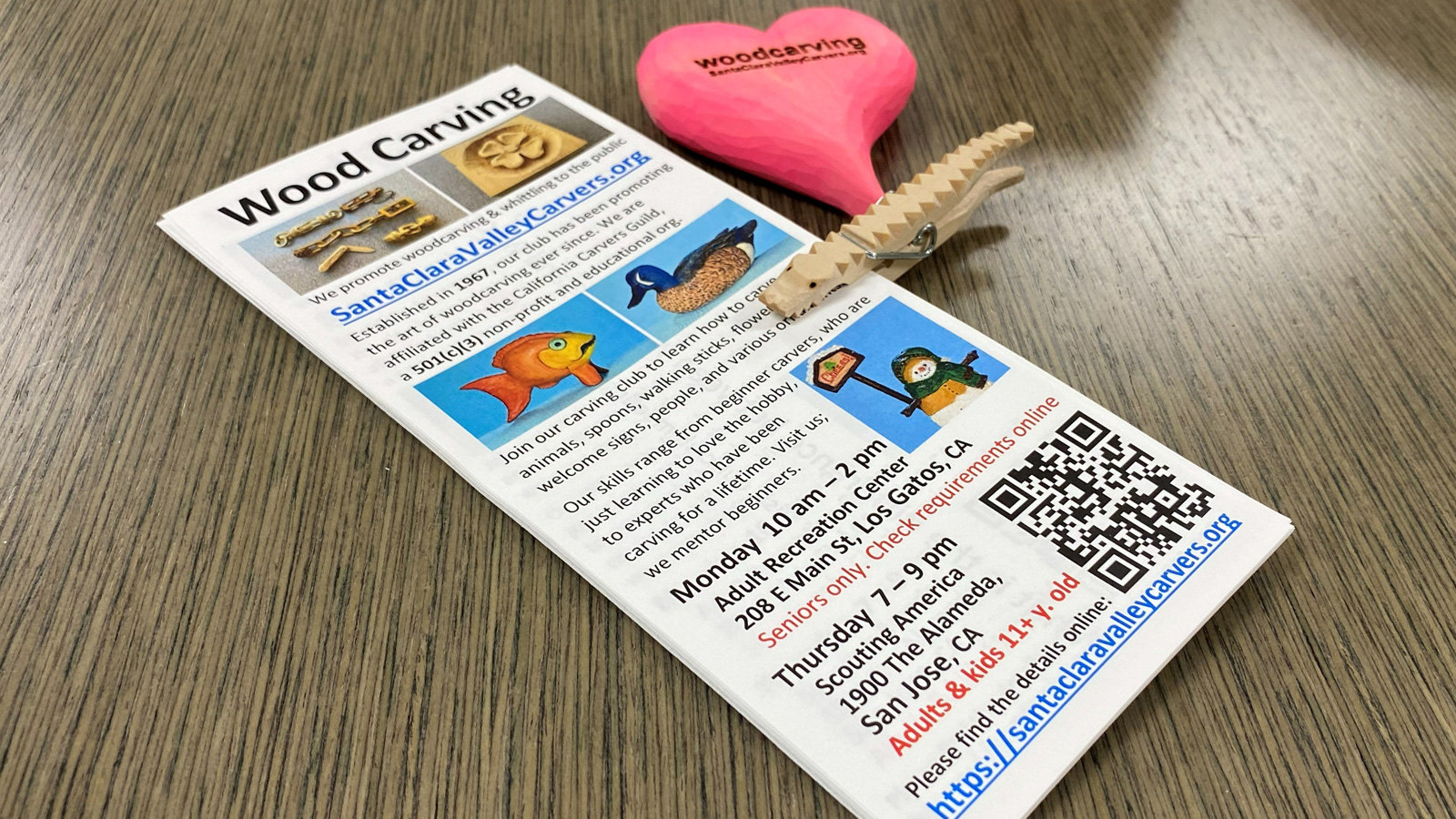 A stack of promotional flyers for "Wood Carving" by the Santa Clara Valley Carvers rests on a table. The woodcarving flyer features photos of carved wooden animals, such as a duck and a fish, along with meeting schedules for Los Gatos and San Jose. Clipped to the top right of the paper stack is a wooden clothespin carved in the shape of an alligator and a bright pink wooden heart engraved with the club's name and website.