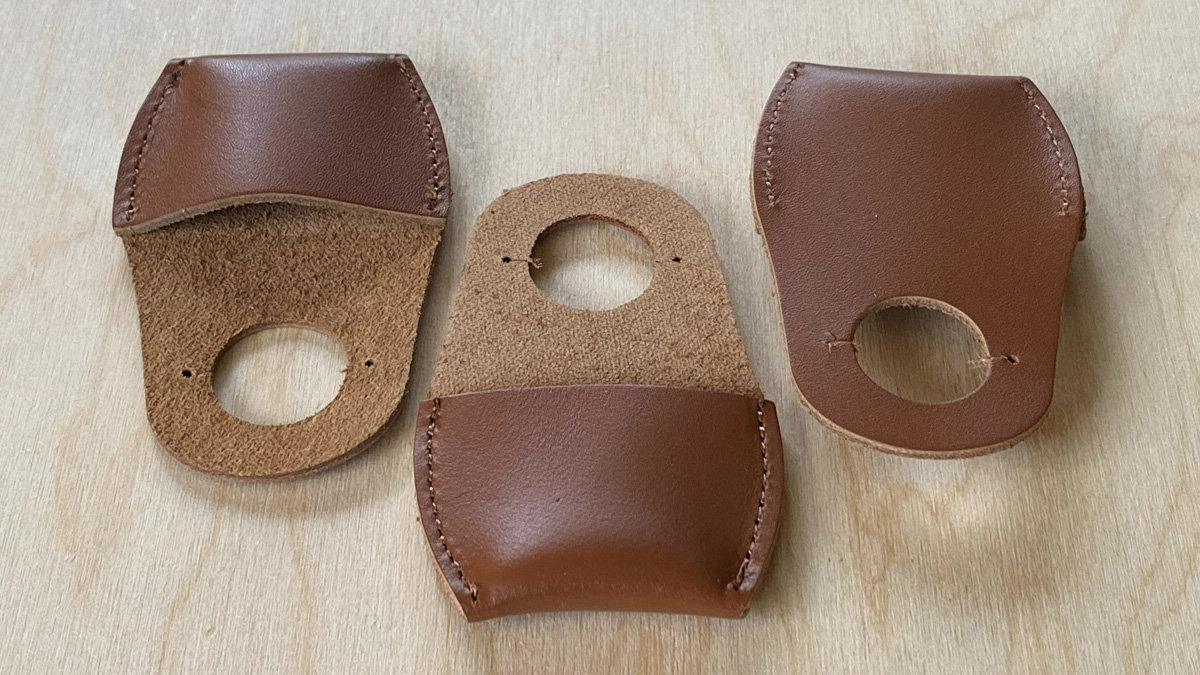 Three brown leather thumb guards used for woodcarving are arranged on a light wooden surface. The guard on the left displays its underside, revealing a rougher, suede-like texture with a circular cutout near the bottom edge flanked by two small punched holes; a smooth leather flap is folded over the top and stitched down. The middle item shows a similar construction but oriented differently, featuring the suede section with the hole at the top and a smooth leather base at the bottom. The guard on the right presents its smooth brown leather exterior, with visible hand-stitching along the side curve and a circular opening near the bottom edge.