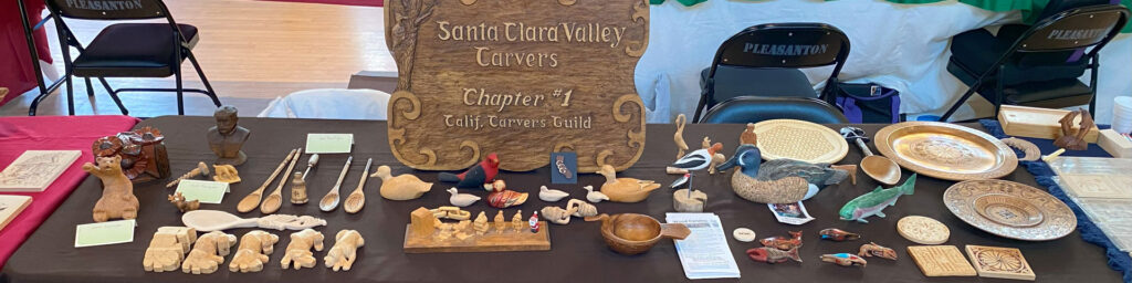 A table set up by Santa Clara Valley Carvers during the Tri-Valley Carvers woodcarving show in 2025. It features carved ducks, spoons, ornaments, and chip-carved coasters.