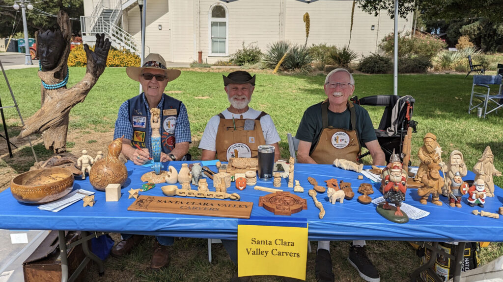 A woodcarving booth with a table full of various wood carving and whittling projects. Three club members presenting the art of whittling and wood carving.