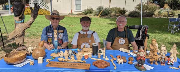 A woodcarving booth with a table full of various wood carving and whittling projects. Three club members presenting the art of whittling and wood carving.