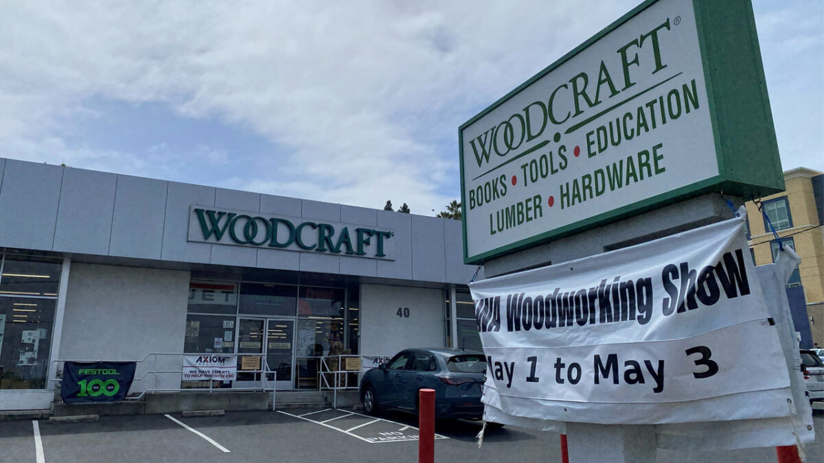 Exterior view of the Woodcraft store in San Carlos, CA, hosting the Bay Area Woodworkers Association Woodworking Show in 2026. A large monument sign reads "WOODCRAFT" with the subtext "Books • Tools • Education" and "Lumber • Hardware." A white banner attached to the signpost announces the "Woodworking Show" dates, May 1 to May 3.