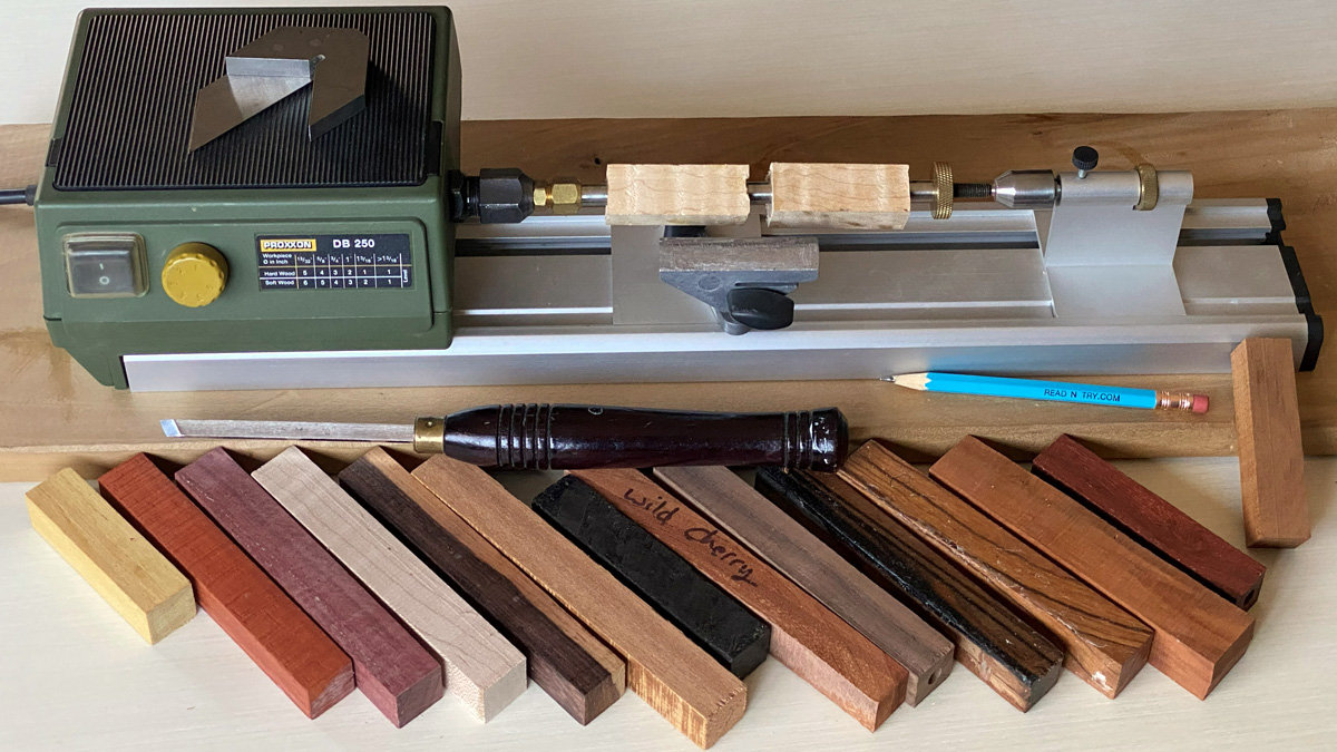 A miniature green Proxxon DB 250 lathe sits at the top of the frame, holding two small blocks of unfinished light wood (maple) on a pen mandrel. Lying horizontally across the metal bed are a woodturning skew chisel with a dark handle and a blue pencil. In the foreground, an assortment of roughly a dozen rectangular wood blanks in various colors, ranging from pale yellow and red to deep browns and black, are arranged diagonally; one of the darker pieces is labeled "Wild Cherry" in handwritten pencil.