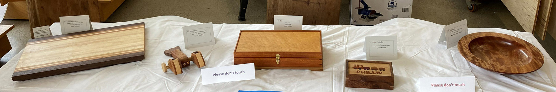A display of custom woodworking projects arranged in a row on a white tablecloth. From left to right, the items include: a long rectangular bread board with a dark wood frame and light wood center panels; a small, striped wooden lobster toy with wheels; a medium-sized lidded box featuring a brass latch and a figured wood top; a small keepsake box with a train design and the name "PHILLIP" pyrographed on the lid; and a large, polished wooden bowl or platter with a rich reddish-brown grain. Small white cards identify each project.
