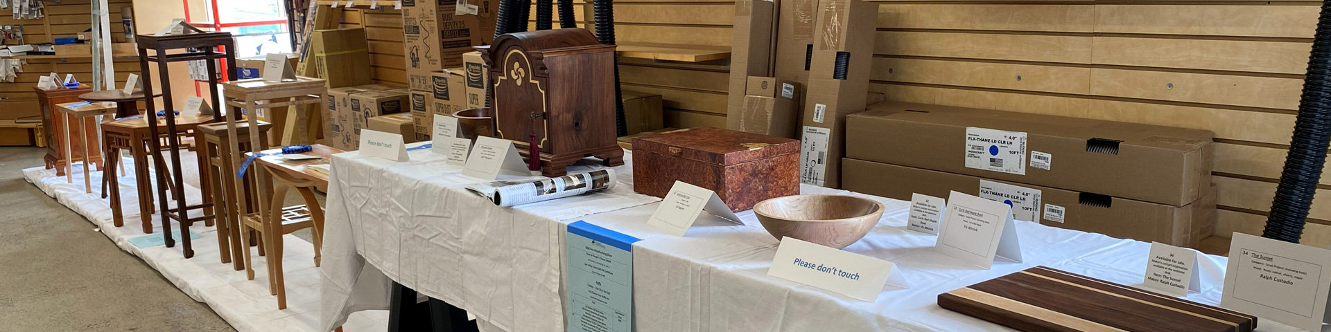 A long table draped in a white cloth displays a variety of handcrafted woodworking projects at a show. On the left, a cluster of wooden stands and small tables includes a tall, dark-wood stand with geometric detailing and a lighter-wood side table. In the center, a dark wood mantle clock featuring light wood inlay and a brass handle sits next to a rectangular box with a mottled, reddish-brown finish. To the right, a large, light-colored wooden bowl is displayed near a striped wooden board in the foreground. White tent cards with project descriptions are placed in front of the items.
