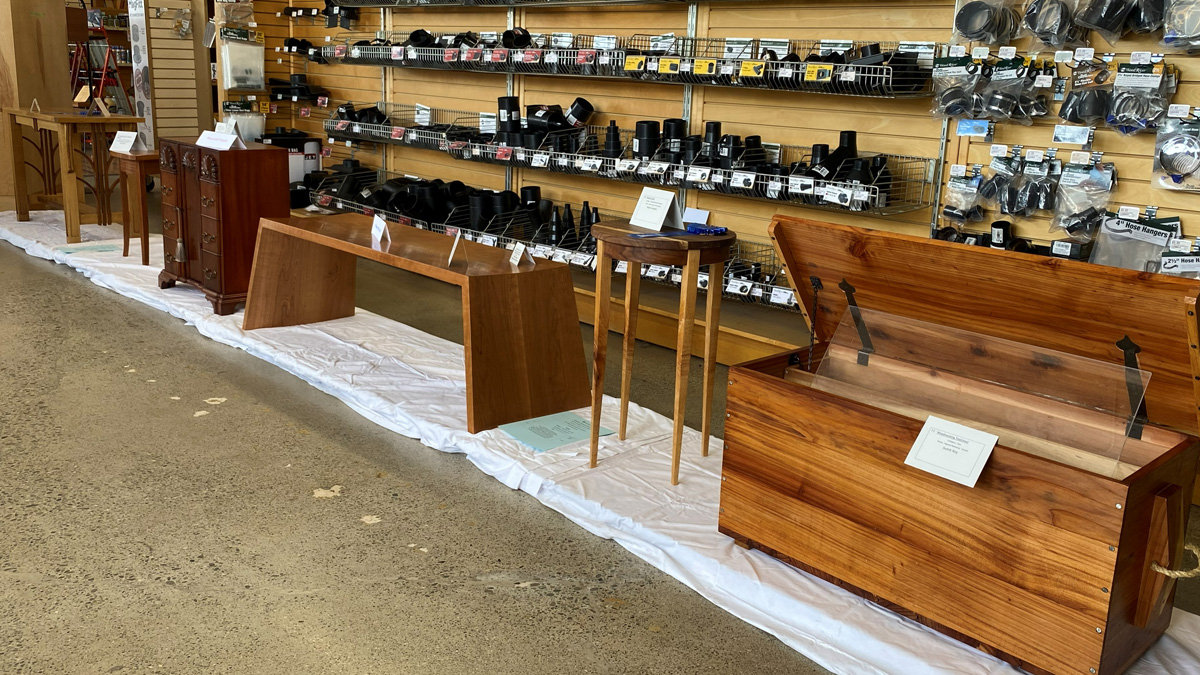 A display of handmade woodworking projects lined up on a white drop cloth on the floor at a woodworking show at Woodcraft store in San Carlos, CA. The pieces include a tall, narrow wooden chest of drawers with brass handles; a long, low bench with a modern design and thick, angled legs; a tall, slender side table with thin, tapered legs; and a large wooden tool chest with rope handles, its lid open. Small white cards with descriptions are placed on or near each project.